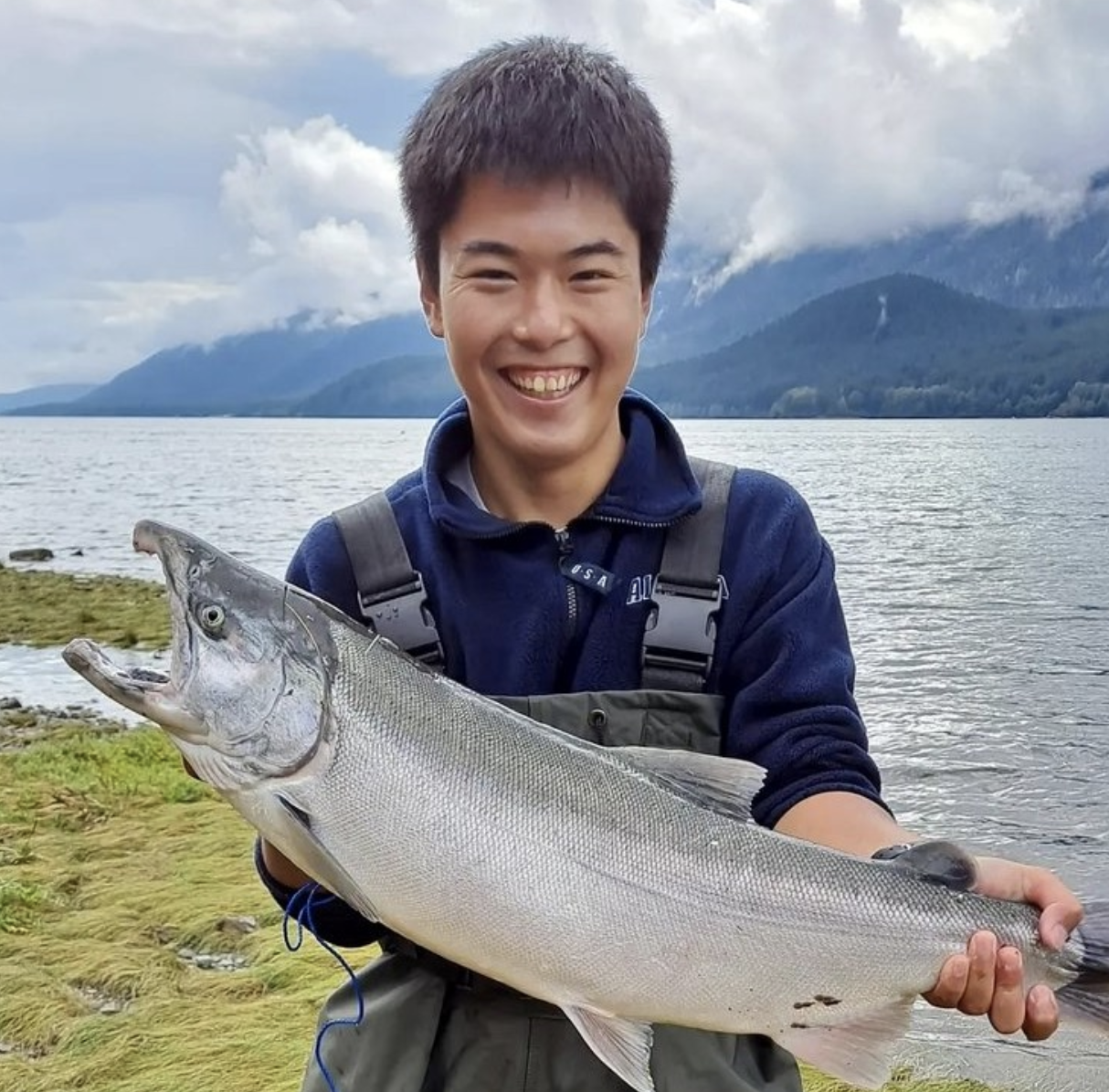 Young person holding a fish and smiling, water and mountain in background.