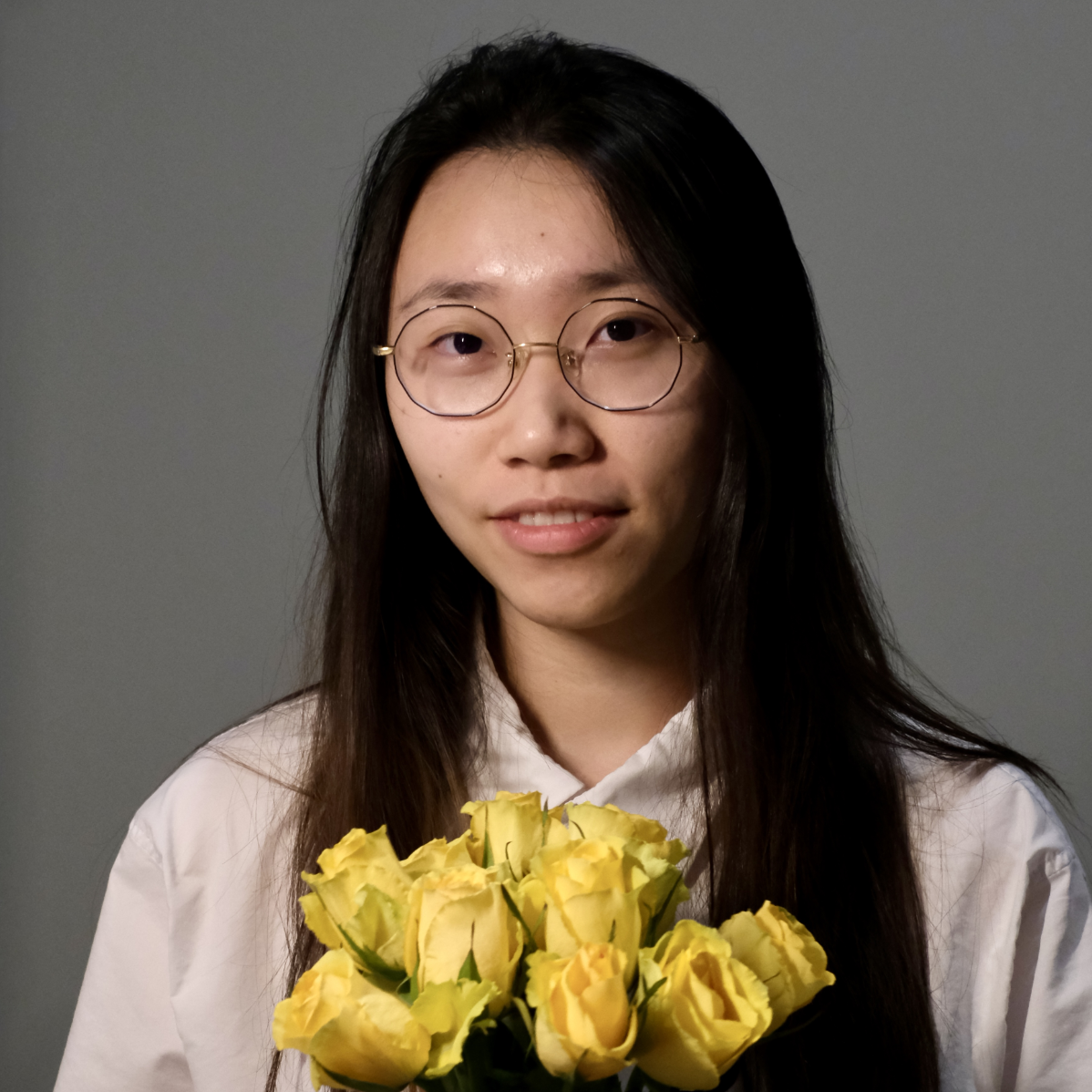Portrait of young woman with long black hair, wire rimmed glasses smiling at viewer and holding a bouquet of yellow roses.