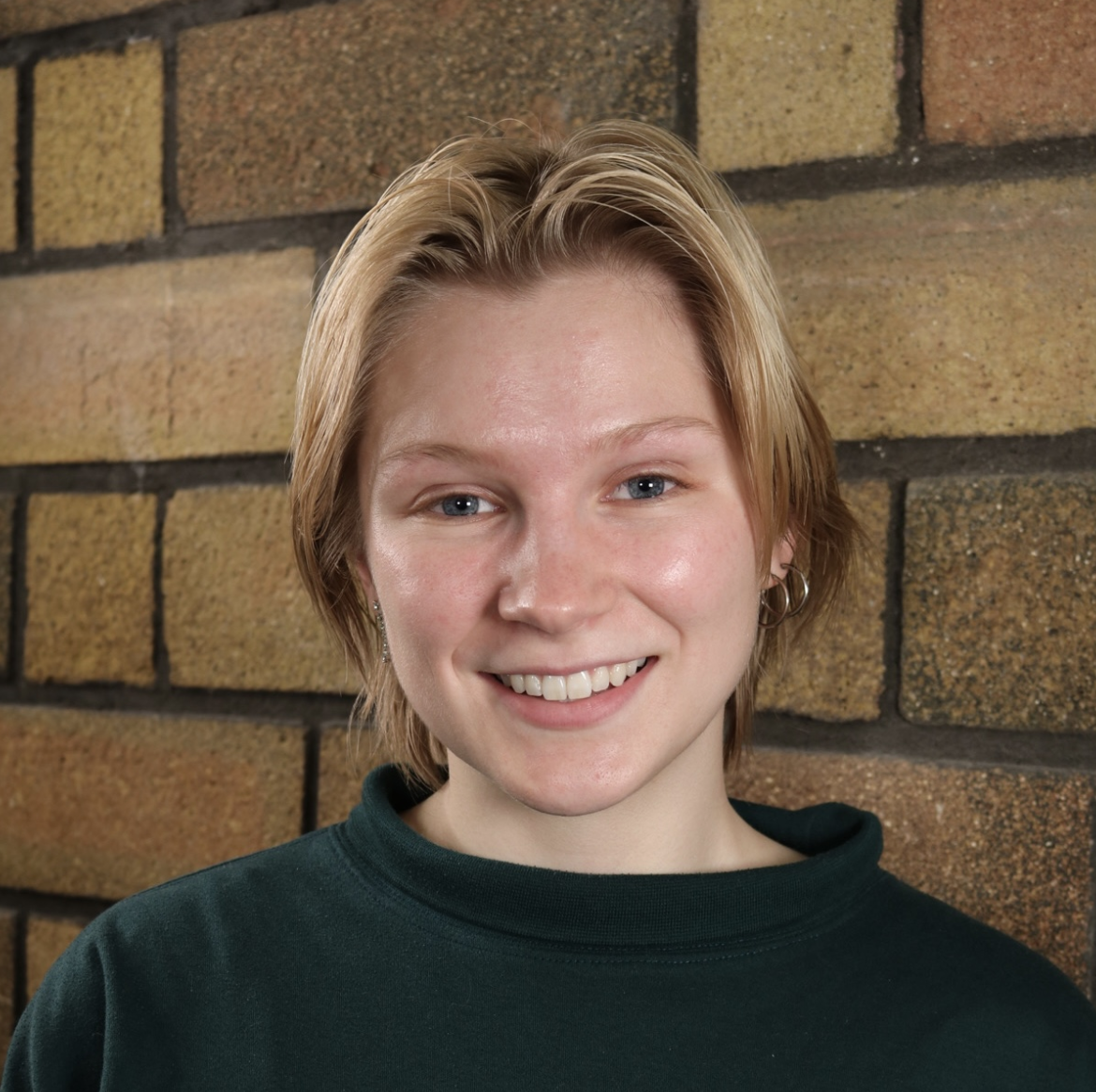 Portrait of young person with short blond hair and green sweater smiling at viewer. Brick wall in background.