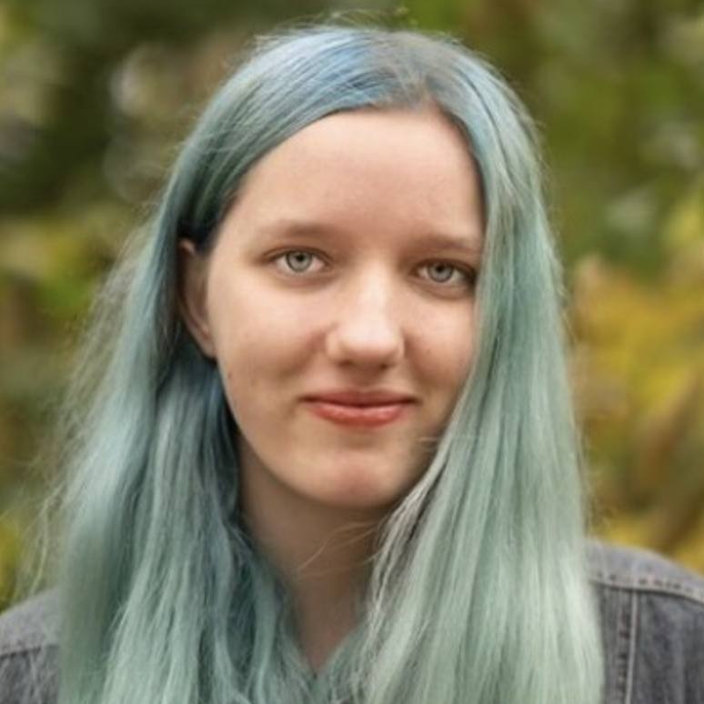 A young person with light teal hair standing outdoors smiles at the camera.