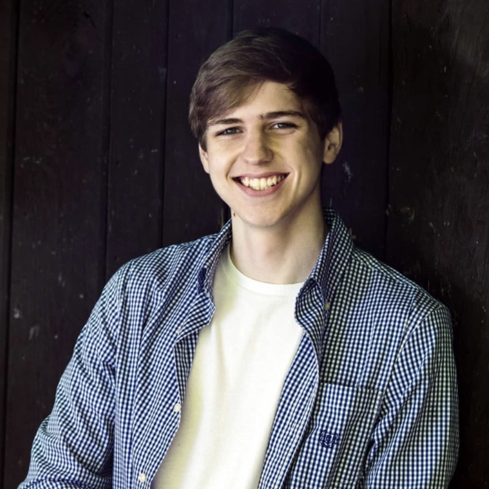 A young person in a blue shirt stands against a dark background and smiles at the camera.
