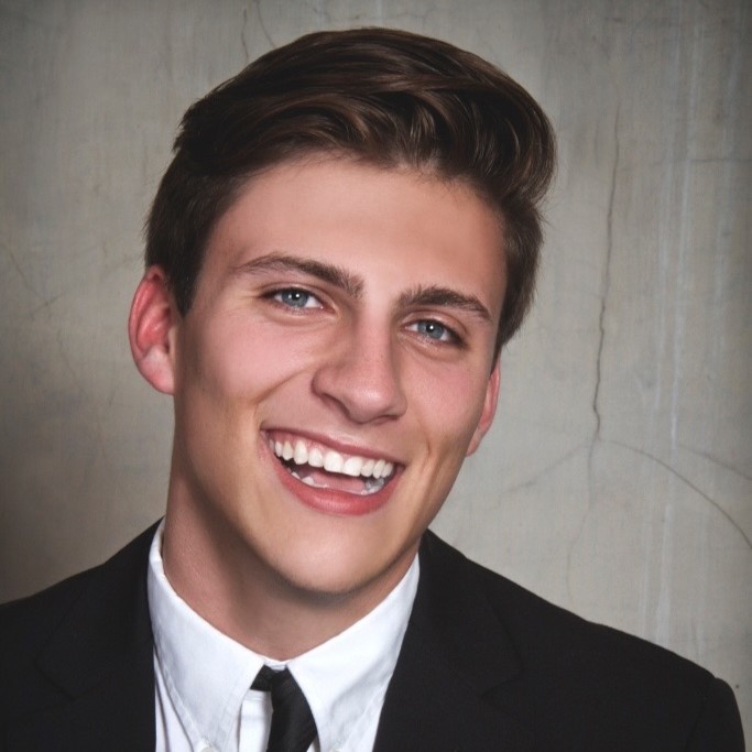 A smiling young man with dark hair wearing a button up white shirt with a black tie and suit jacket in front of a gray wall