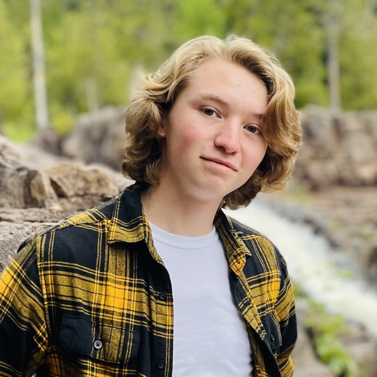 A young man in front of a stream with curly blond hair wearing a white tee with a yellow and black plaid shirt on top