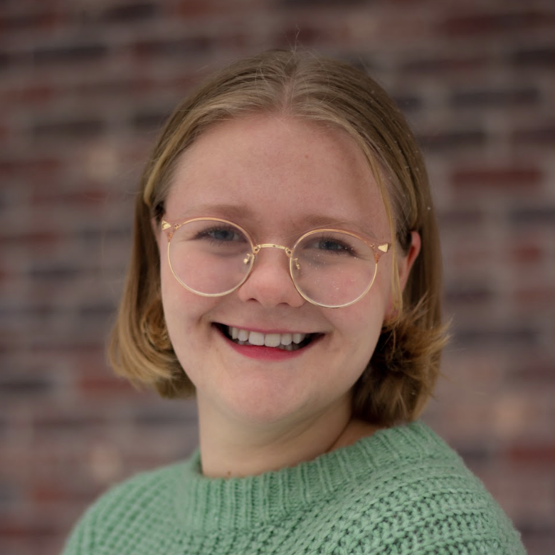 A young person wearing glasses and a green sweater smiles.