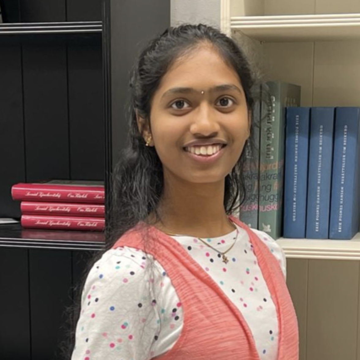 A young person in a polka dot shirt stands near a bookshelf and smiles.