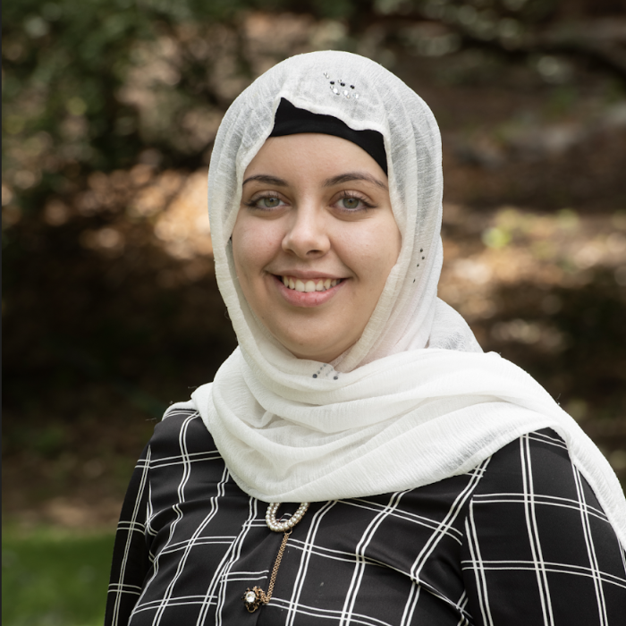 A young person in a plaid shirt and white hijab smiles at the camera.