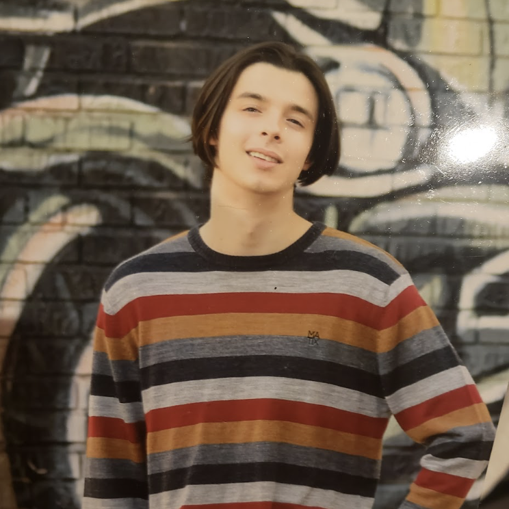 A young person in a striped sweater stands near a wall and smiles.