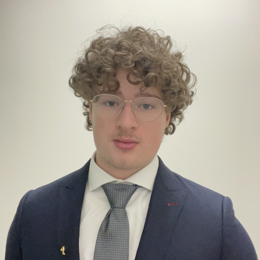 A male-presenting person with curly hair and glasses wears a suit.