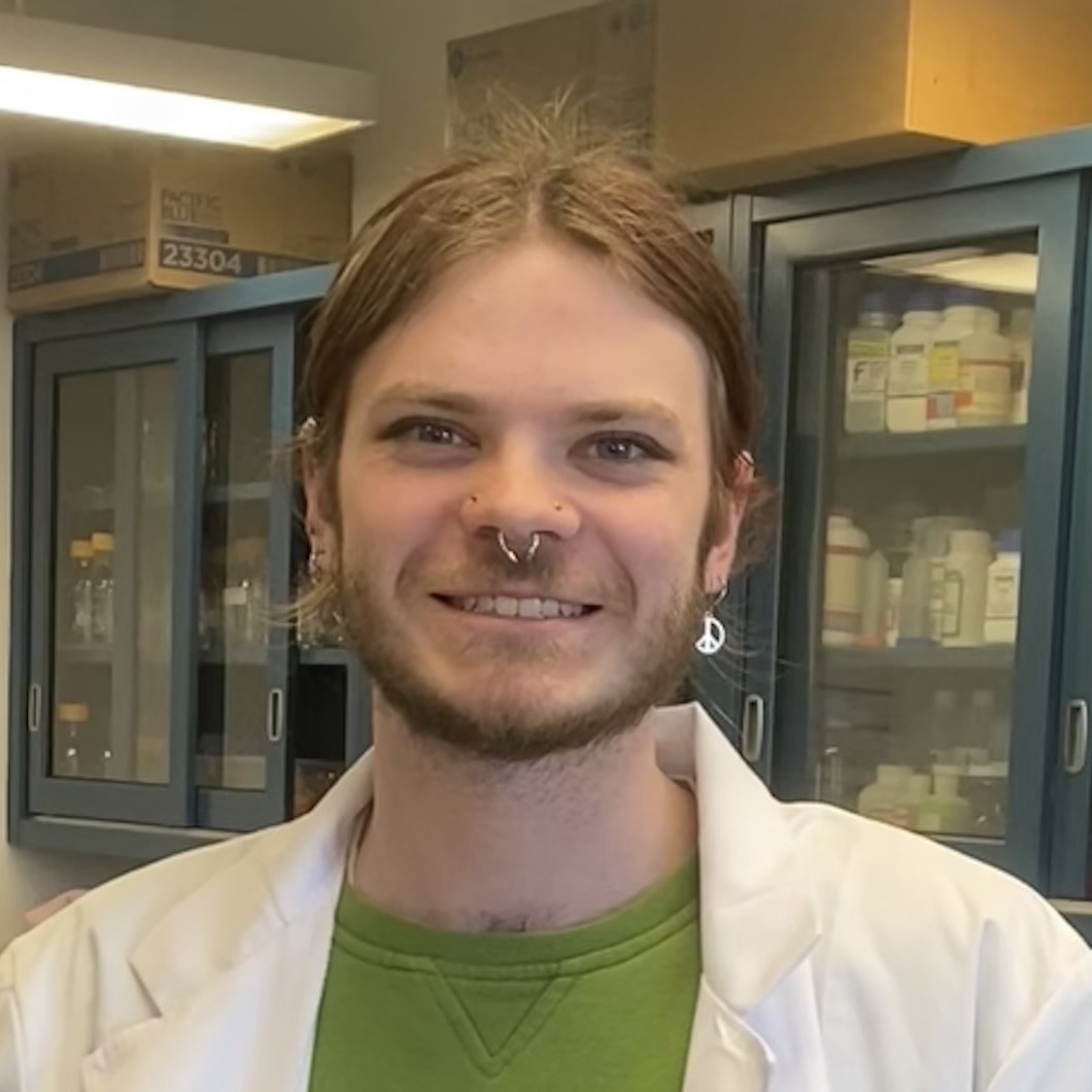a young person with facial hair with a nose and ear piercing wearing a lab coat smiling